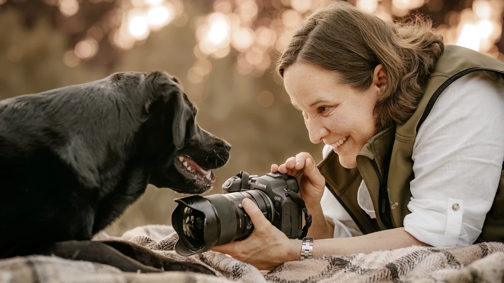Eine Frau mit einer Kamera lächelt und fotografiert einen schwarzen Labrador, der ihr gegenüber liegt. Sie befinden sich im Freien auf einer Decke.