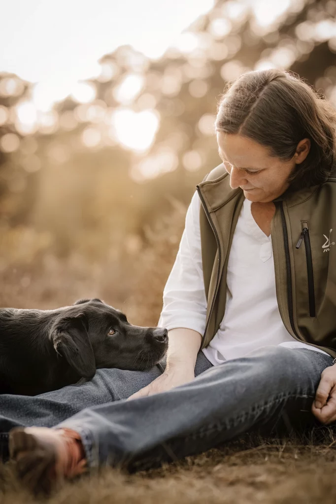 Hundefotografin Silke Schmitt aus Münster genießt einen ruhigen Moment in der Abenddämmerung mit ihrer schwarzen Labrador Hündin Pepper, die ihren Kopf liebevoll auf ihrem Bein abgelegt hat.