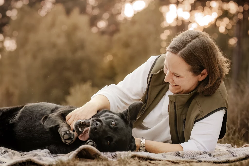 Eine Frau liegt auf einer Decke und spielt mit ihrem schwarzen Labrador, der sich auf dem Rücken wälzt. Beide genießen einen fröhlichen Moment in der warmen Abendstimmung.
