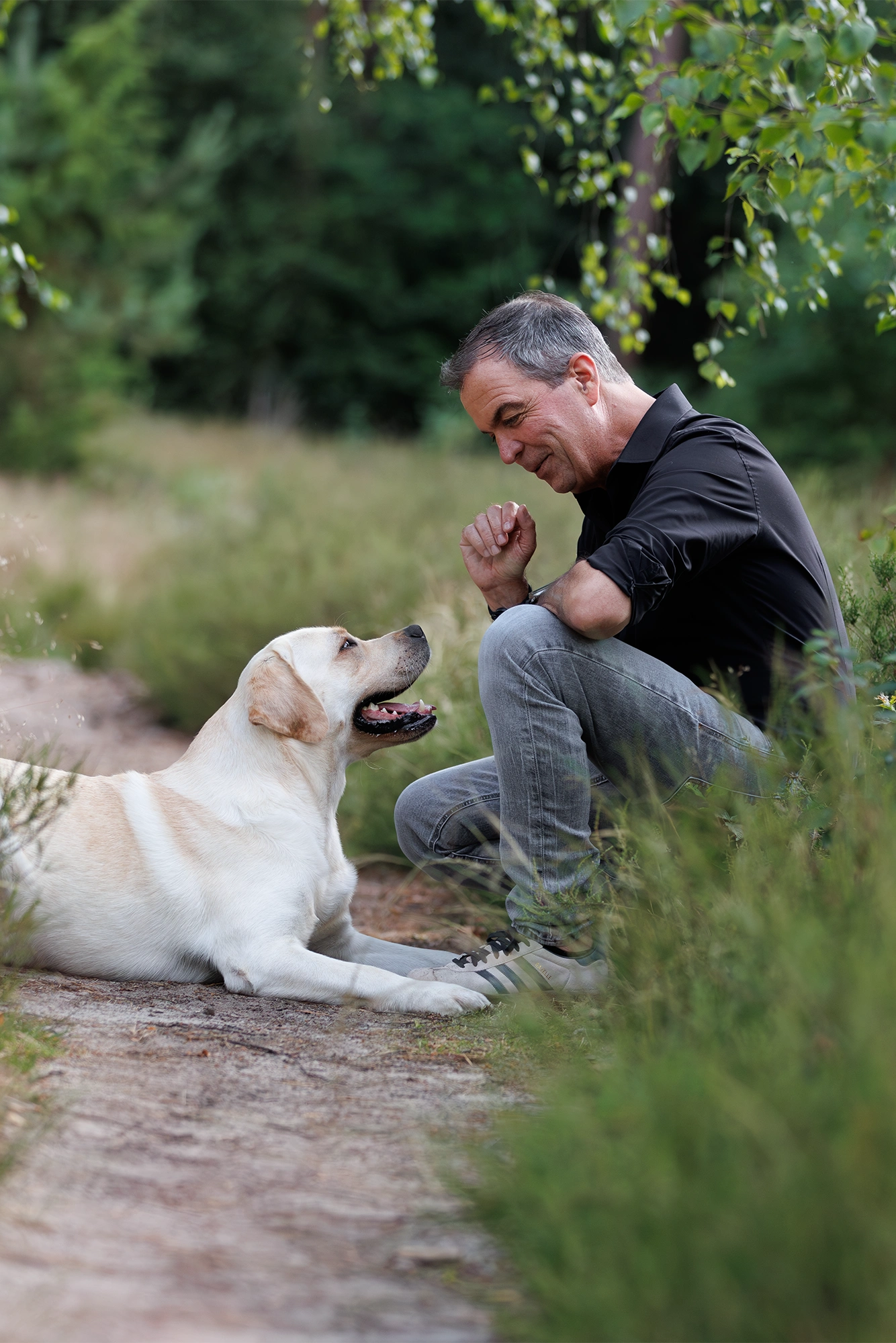 Peter kniet neben seinem Labrador Theo in einem Wald, während sie sich freundlich anschauen. Der Hund liegt entspannt auf dem Boden.