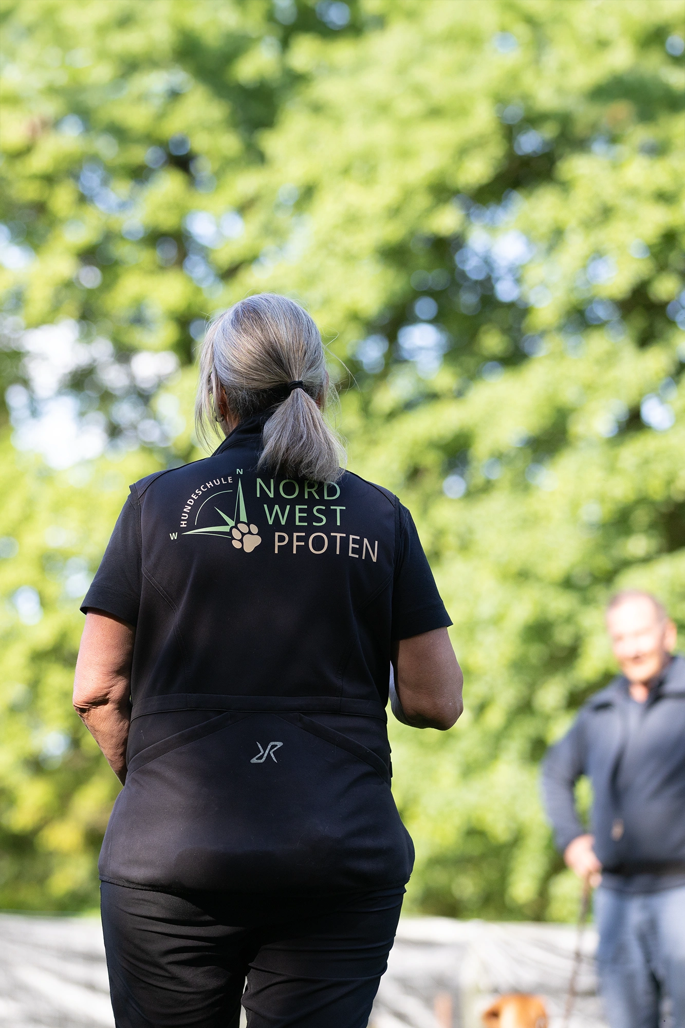 Eine Person mit grauen Haaren trägt ein schwarzes T-Shirt mit dem Aufdruck "NORD WEST PFOTEN". Im Hintergrund sind grüne Bäume und eine weitere Person mit einem Hund zu sehen.